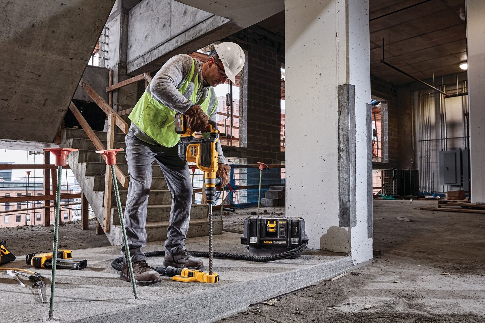 Worker using DEWALT power tool for drilling concrete on construction site; equipment and toolbox are nearby.